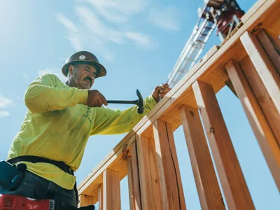 A carpenter in hi-vis nails timber studs into a wall frame, with a tower crane in the sky behind